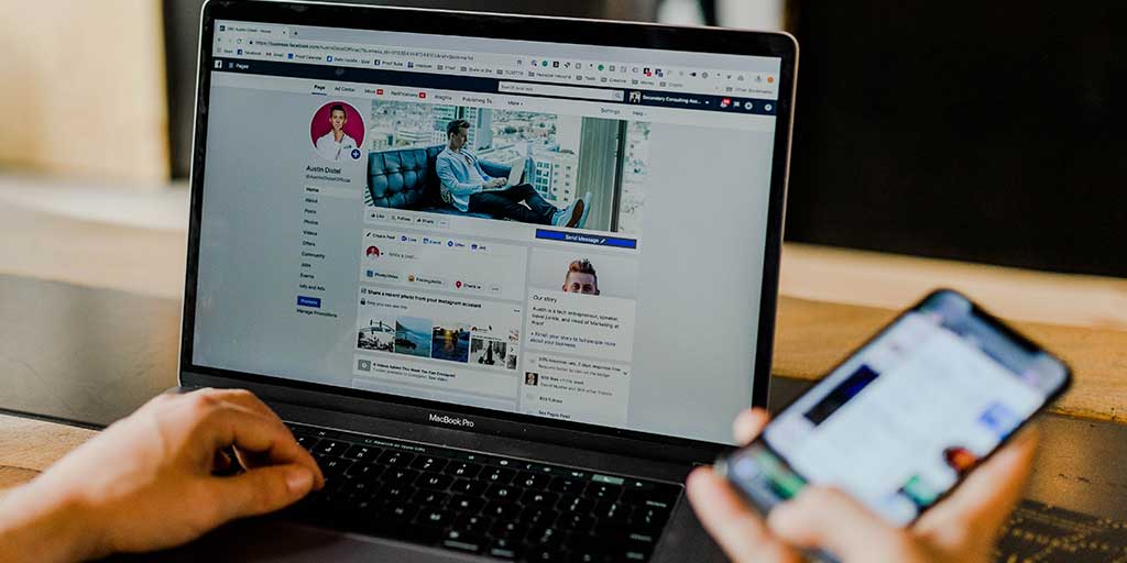 uma mão segurando um celular e outra digitando no teclado de notebook acessando um perfil no Facebook
